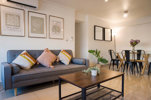 Una sala de estar moderna con un sofá gris, una mesa de centro y un comedor con mesa y sillas. Hay obras de arte enmarcadas en la pared y una planta en maceta sobre la mesa de centro.