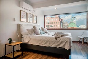 A bedroom with a bed, a nightstand, a lamp, a chair, and a window with a city view.
