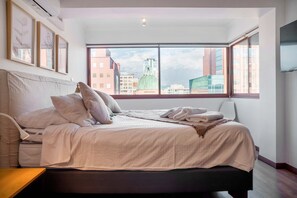 Un dormitorio con un ventanal grande, una cama con almohadas, una mesita de noche y vistas a edificios.