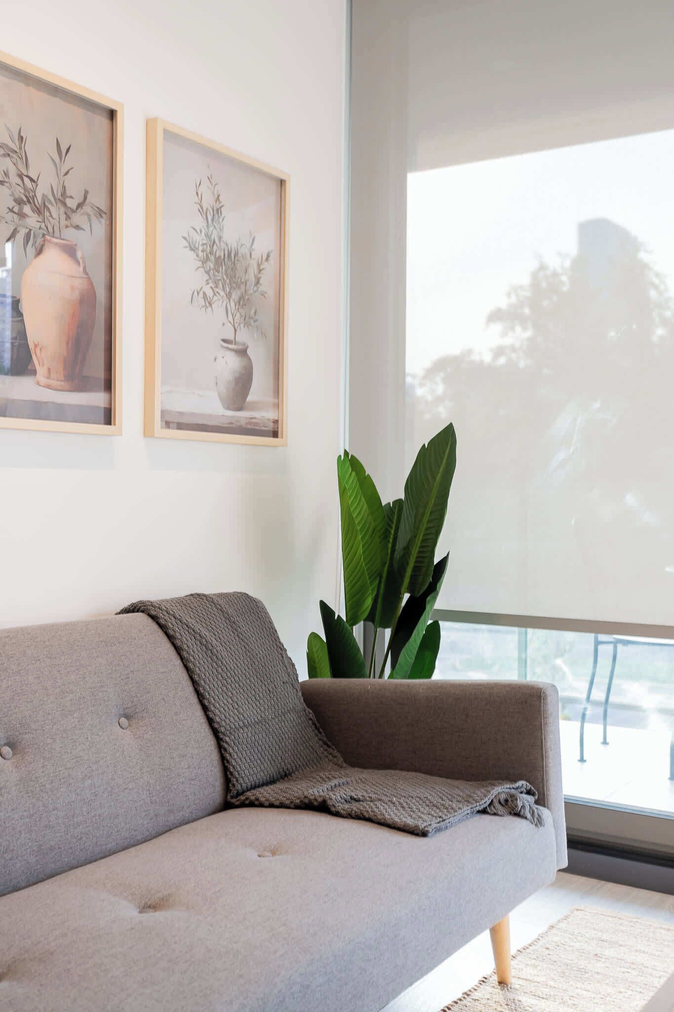 Ein modernes Wohnzimmer mit grauem Sofa, einem großen Fenster und zwei gerahmten Pflanzenbildern an der Wand.