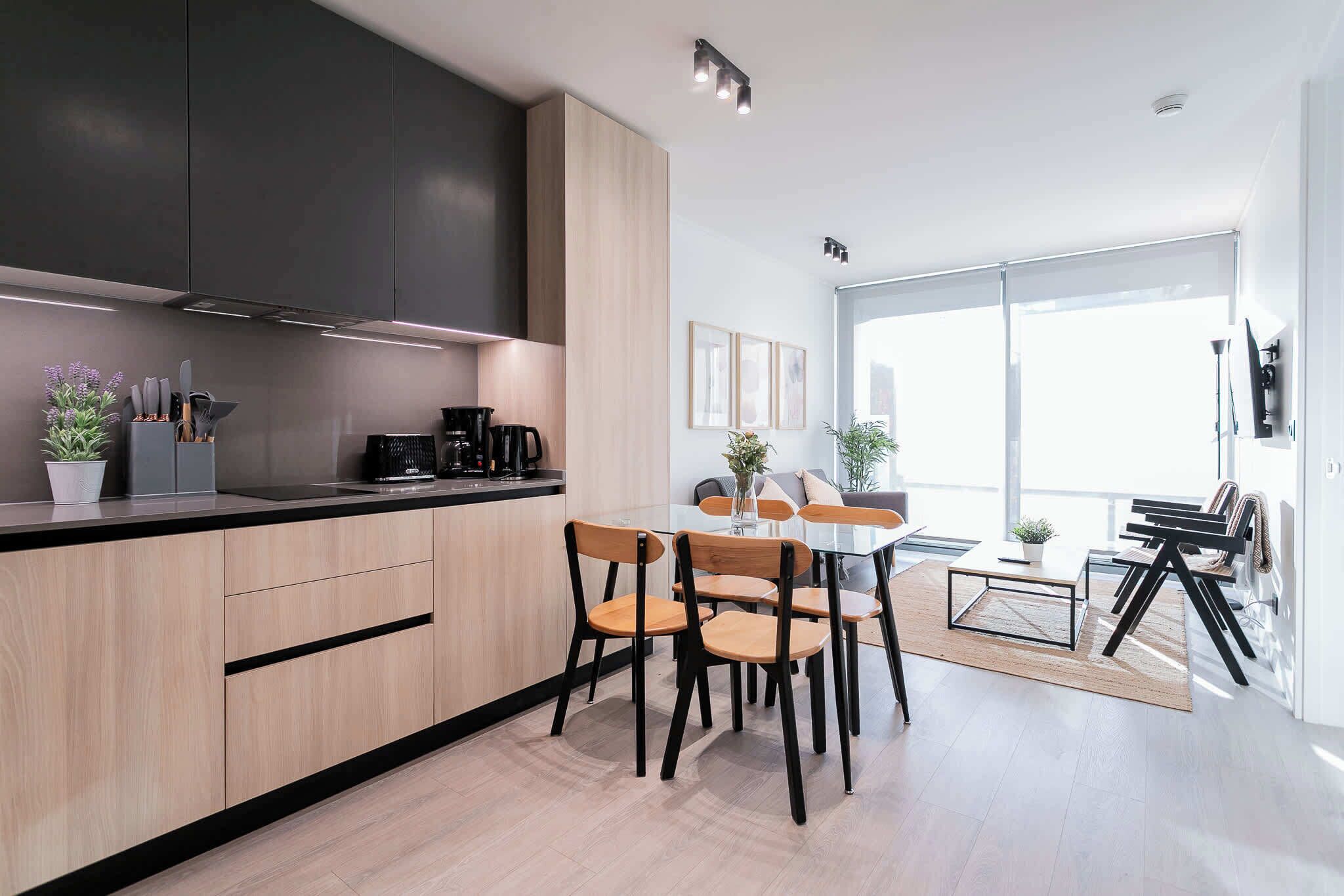 Een moderne keuken met houten kastjes, een eethoek met een glazen tafel en stoelen, en een groot raam dat natuurlijk licht binnenlaat.