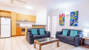 A living room with two sofas, a glass coffee table, and a kitchen area with wooden cabinets and a white refrigerator.