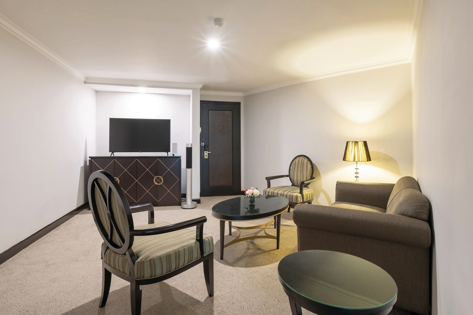 A hotel room with a TV, a sofa, a chair, a small table, and a lamp.