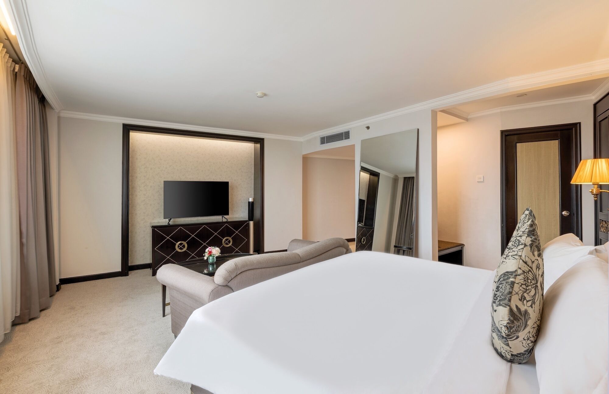 A hotel room with a bed, a sofa, a TV on a stand, a mirror, and a lamp.