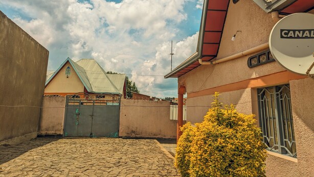A courtyard with a cobblestone floor, a building with a metal roof, and a bush in the foreground. - Rand's House I Homestay (Ruhengeri)