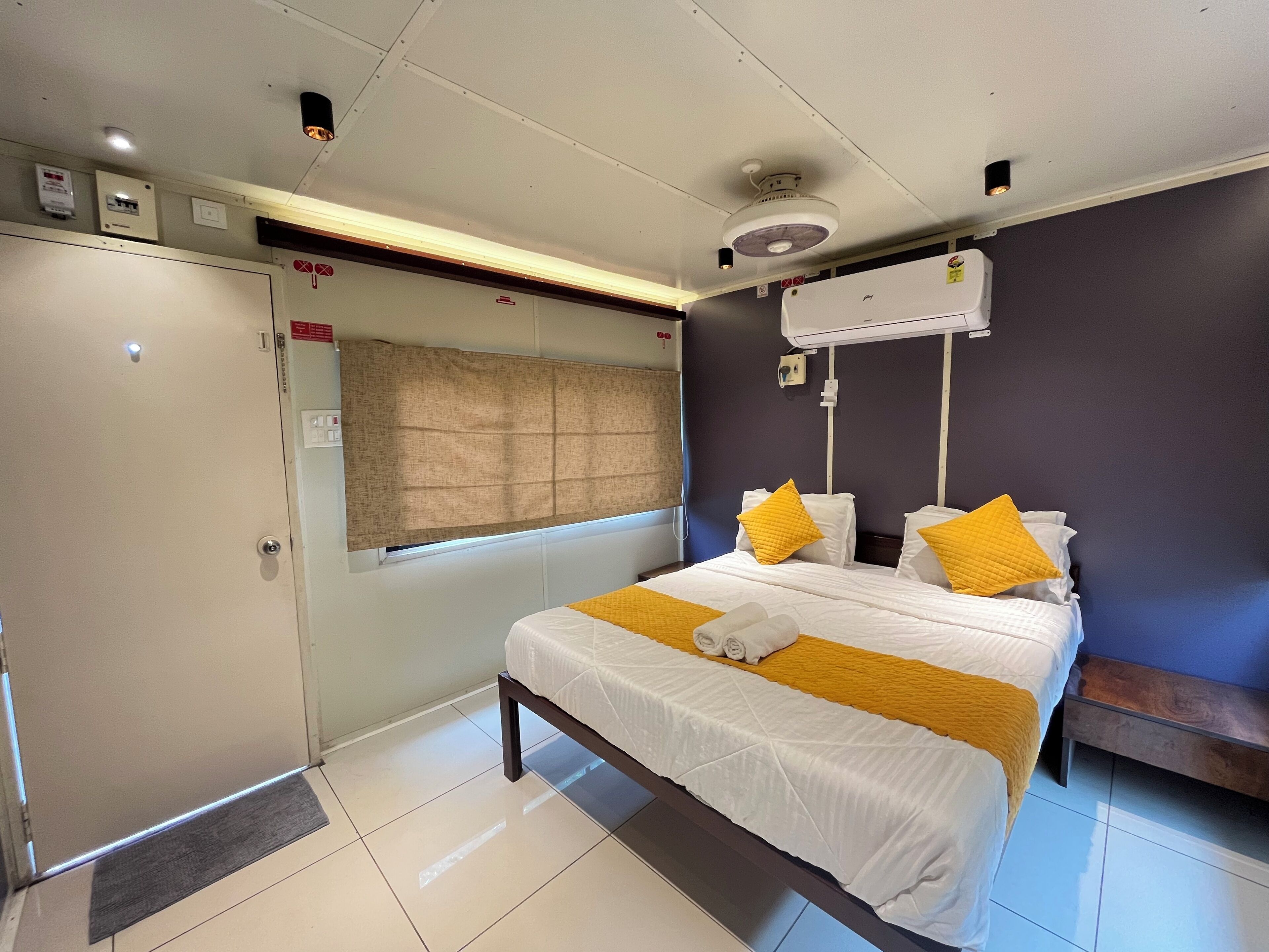 A hotel room with a bed, a bedside table, a window with a curtain, an air conditioning unit, and a door with a handle.