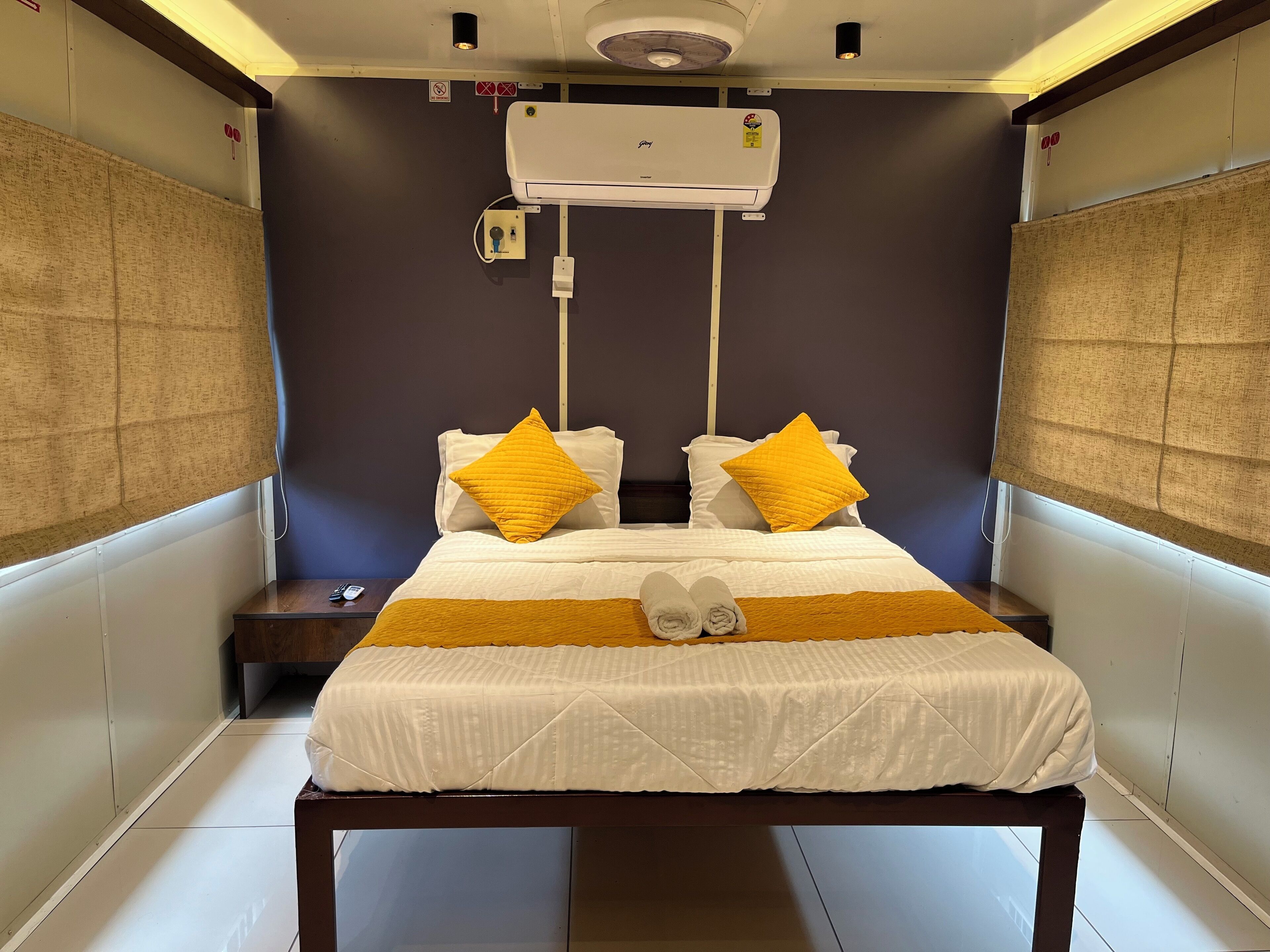 A hotel room with a bed, two pillows, a bedside table, and an air conditioning unit.