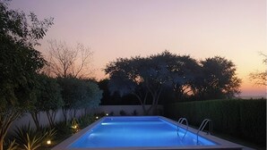 Una piscina rectangular con solárium alicatado, rodeada de setos y árboles bien cuidados, bajo un cielo crepuscular.