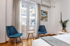 Un dormitorio moderno con una cama, dos sillas azules, una mesita redonda, una planta en maceta y vistas de la ciudad a través de una puerta corredera.
