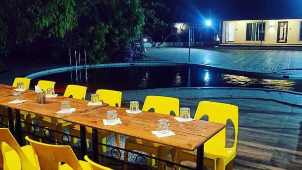 Un espace extérieur aménagé pour les repas, avec une longue table en bois et des chaises jaunes, entouré d’une piscine et d’un bâtiment en arrière-plan.