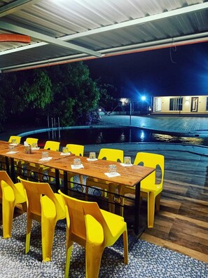 An outdoor dining area with a long wooden table and yellow chairs, surrounded by a pool and a building in the background. - Evergreen Farms (Karjat)