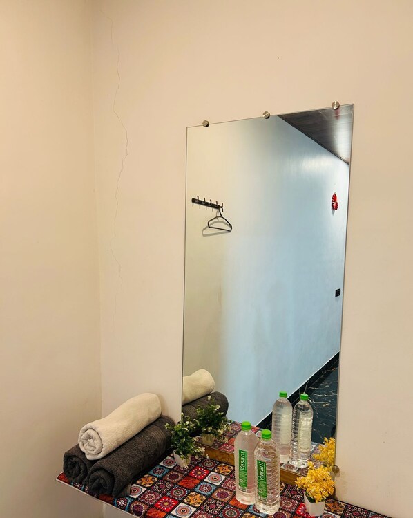 Une salle de bain avec un grand miroir, un porte-serviettes, une étagère avec des serviettes pliées, une petite plante en pot et deux bouteilles d’eau.