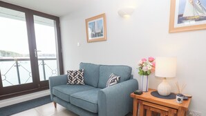 A modern living room with a blue sofa, two patterned cushions, a small wooden side table with a lamp, a vase of flowers, and a framed picture on the wall. - Tobys Quay (Falmouth)