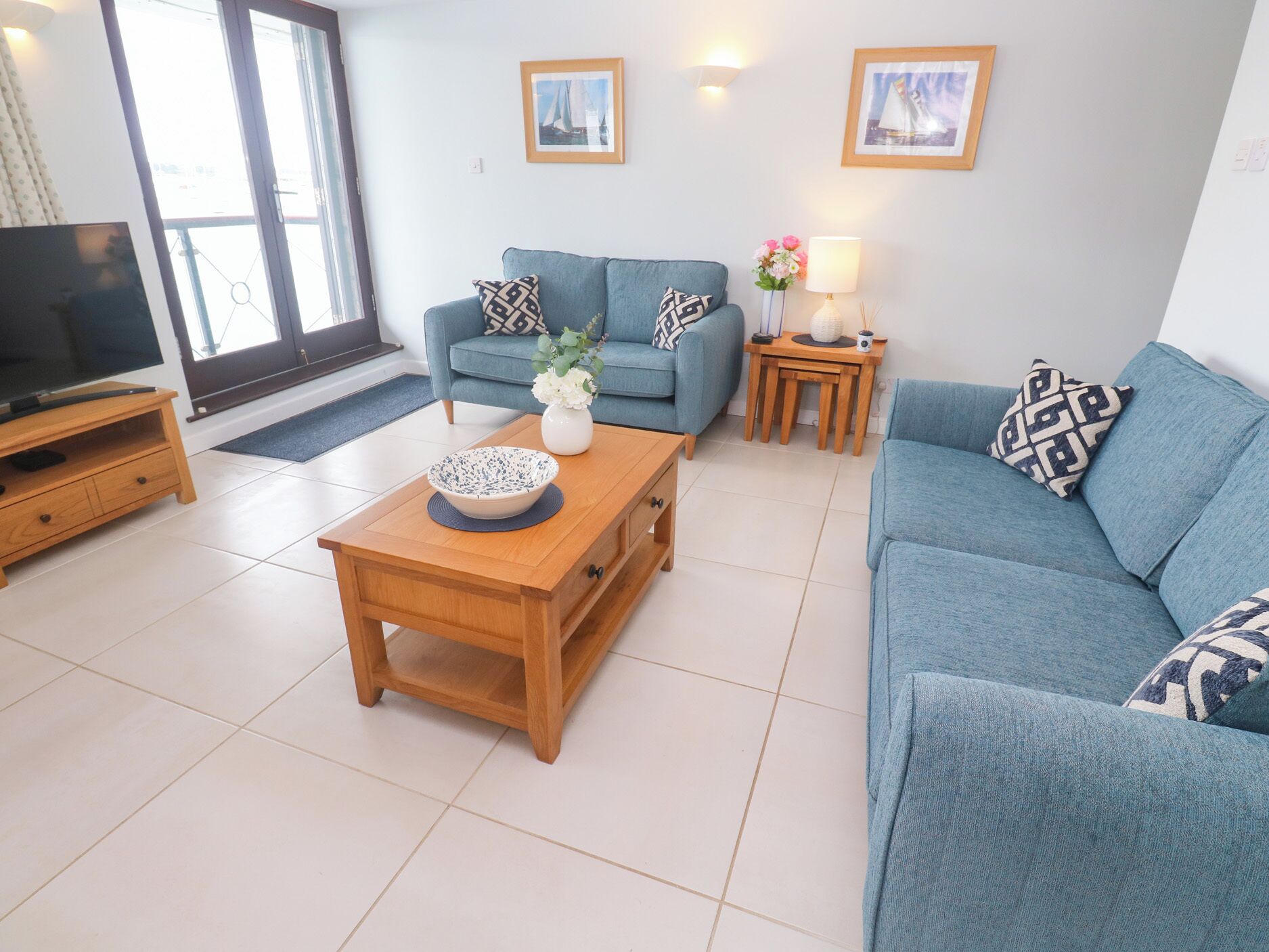 A modern living room with a blue sofa, a wooden coffee table with a bowl, a TV on a wooden stand, and a small side table with a lamp and flowers.