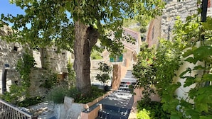 A stone building with a staircase leading up to a door, surrounded by greenery and a tree.