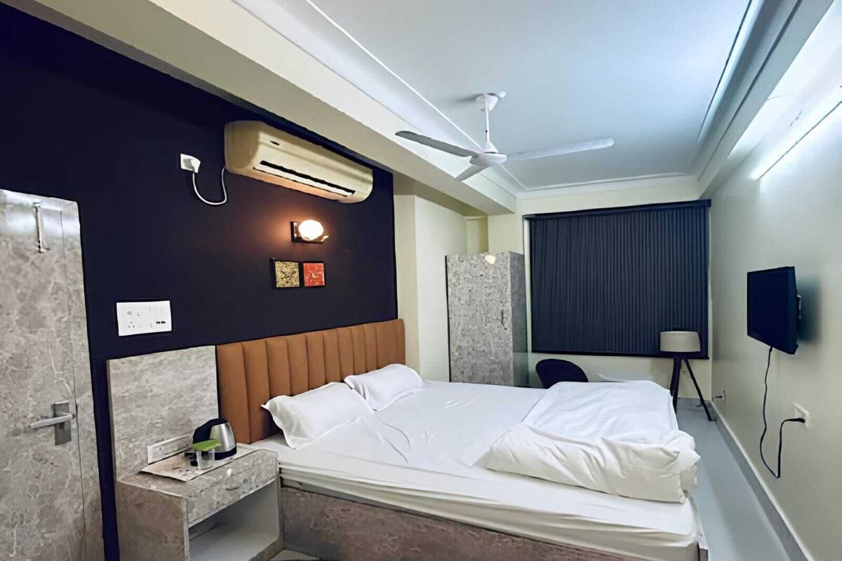 A modern hotel room with a bed, bedside table, air conditioning unit, ceiling fan, and a TV mounted on the wall.