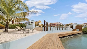 A wooden deck leading to a beach with lounge chairs and a palm tree. - Kas Blou Blou with refreshing communal pool (Kralendijk)