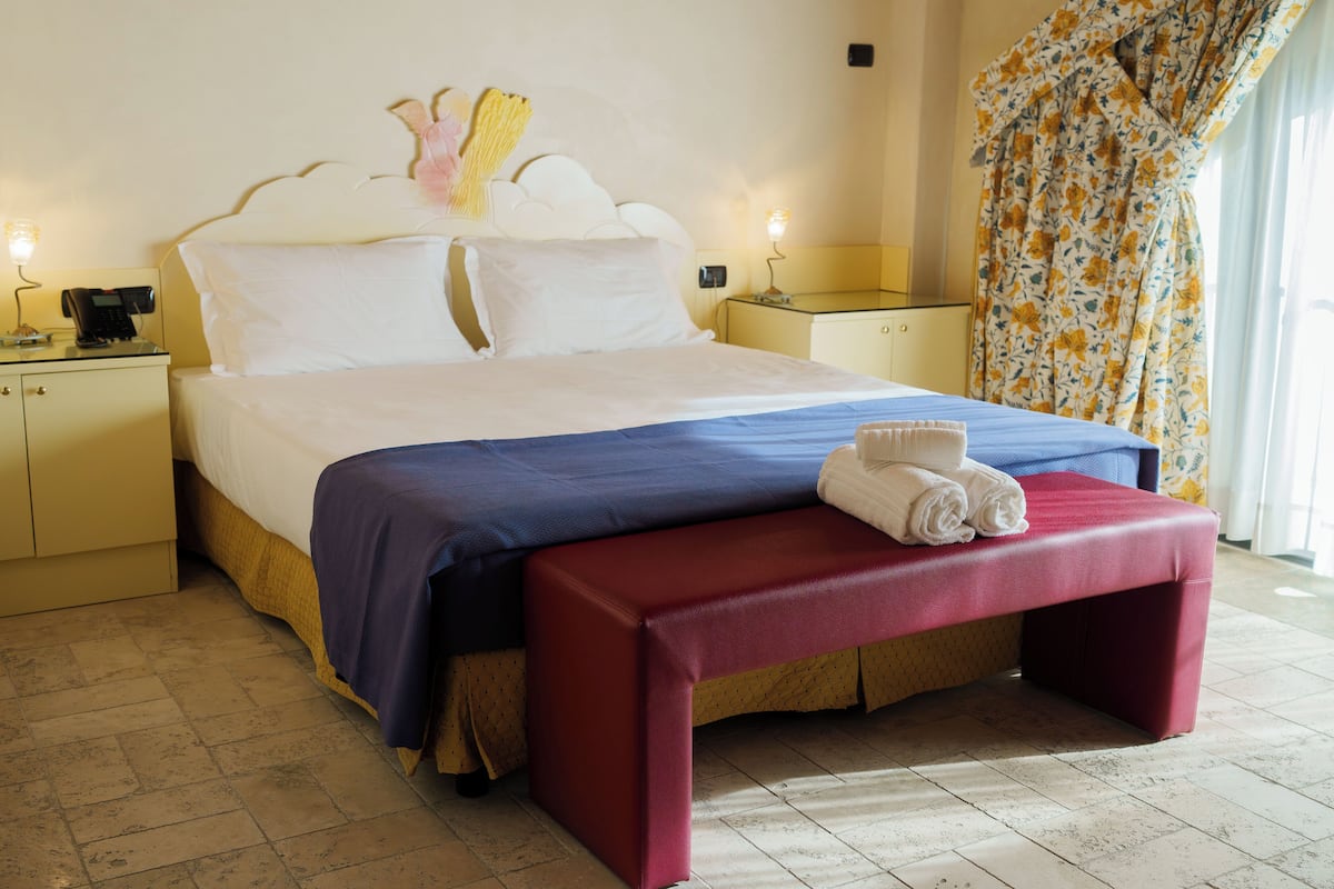 A hotel room with a bed, a bedside table, a nightstand, a bench, and a window with curtains.