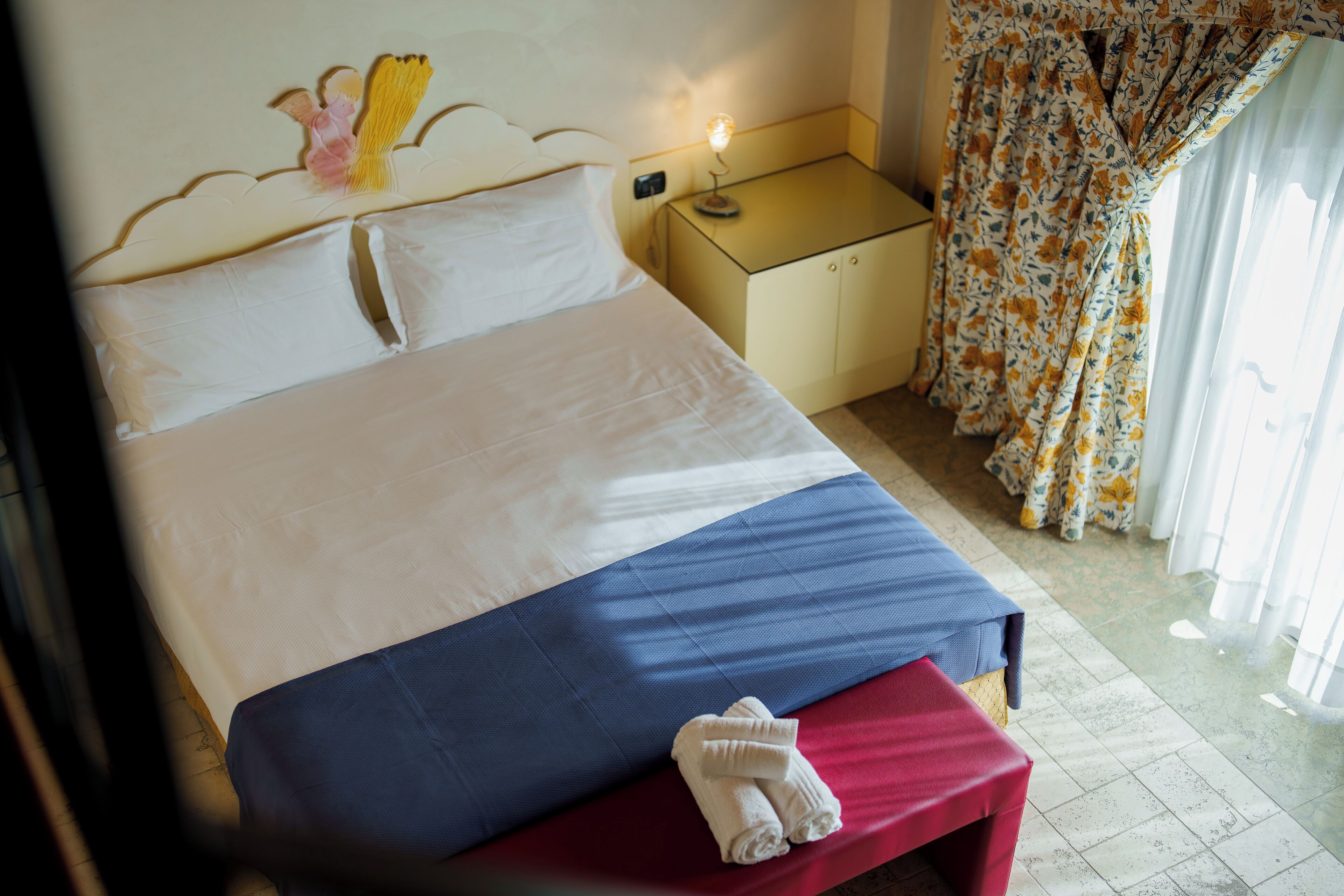 A hotel room with a bed, a bedside table with a lamp, a small bench with towels, and a window with curtains.
