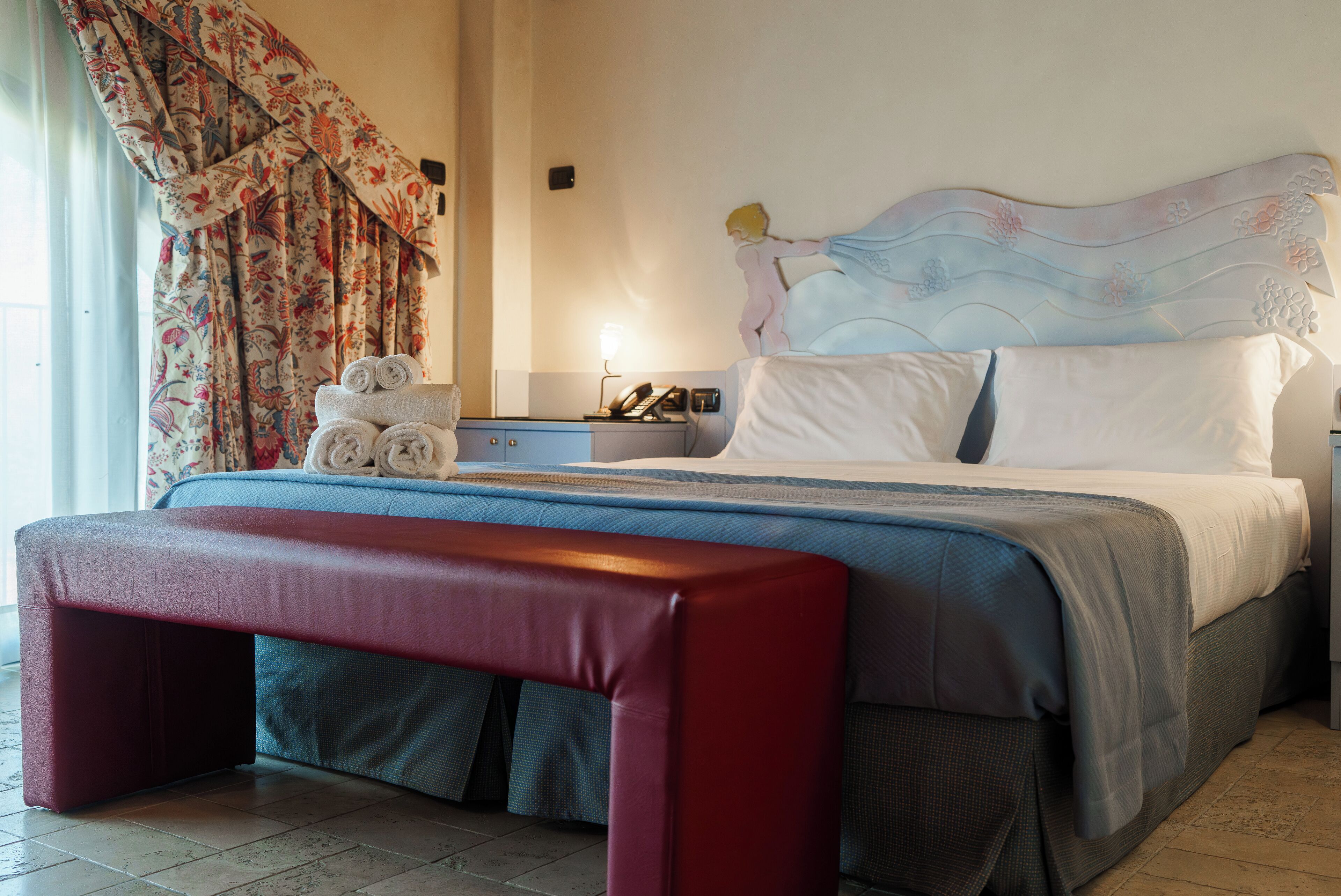 A neatly made bed with a headboard, a bedside table with a lamp, and a bench in front of the bed.
