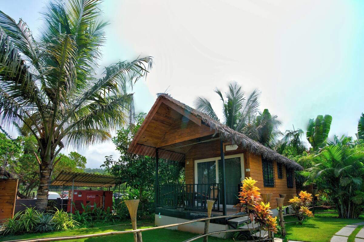 A wooden house with a thatched roof, surrounded by lush greenery and palm trees.