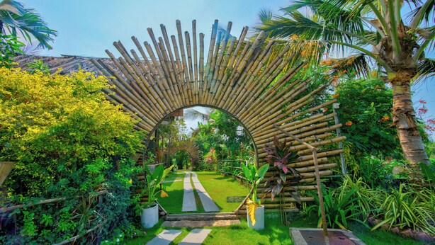 A bamboo archway leading to a garden with a pathway, surrounded by lush greenery and palm trees. - The Lazy Cabanas (Candolim)