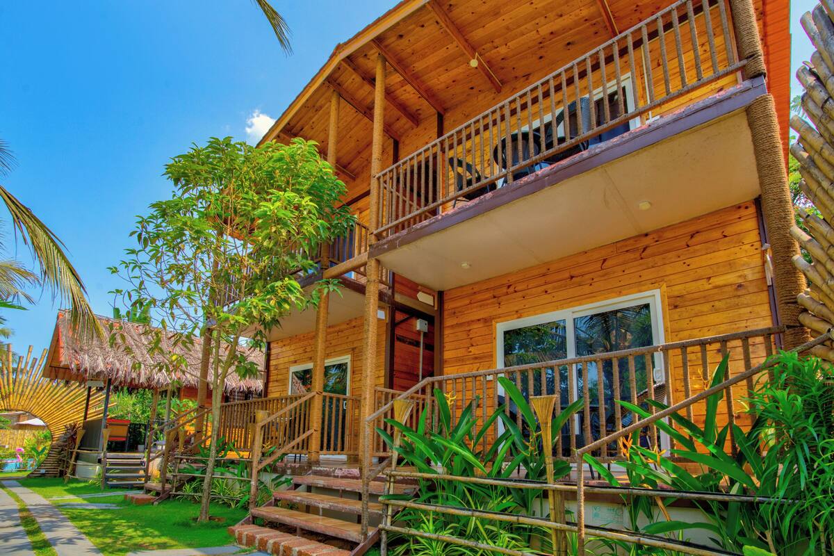 A wooden two-story building with a balcony, surrounded by lush greenery and a clear blue sky.
