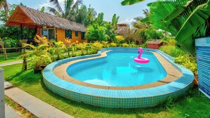 A swimming pool with a tiled edge, surrounded by lush greenery and a thatched-roof structure. - The Lazy Cabanas (Candolim)