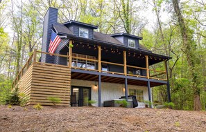 A two-story house with a balcony, a flag, and string lights. - Private Creekfront Cabin on Fightingtown Creek | Hot Tub, Game Room & Fishing (Epworth)