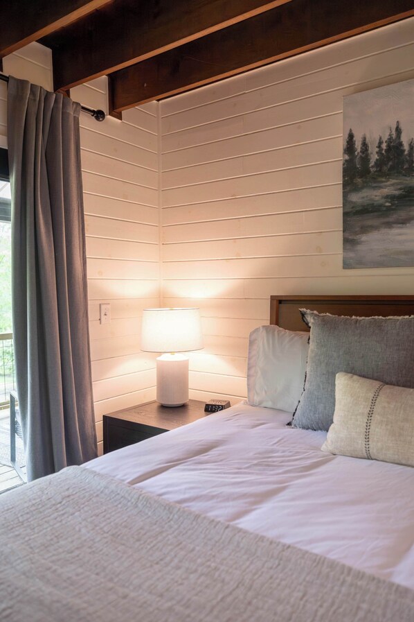 A bedroom with a bed, a nightstand, a lamp, a painting, and a window with curtains.