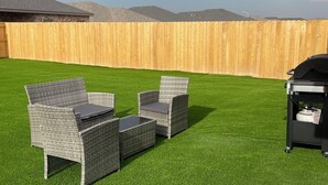 Un patio trasero con césped artificial, mobiliario de mimbre y una parrilla de barbacoa.