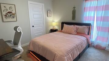 Un dormitorio con cama, escritorio, silla, puerta y ventana con cortinas.
