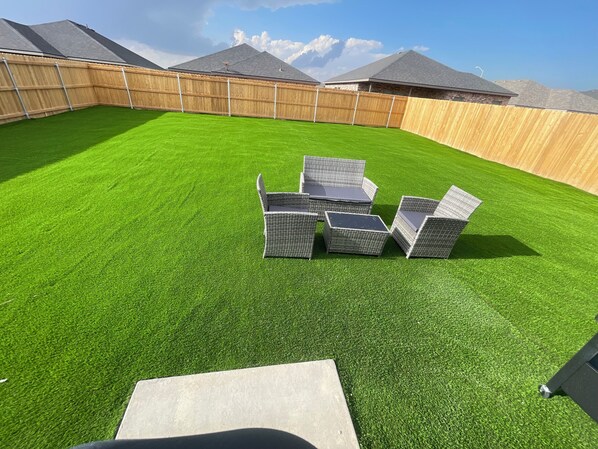 Ein Garten mit künstlichem Rasen, einem Holzzaun und einer Terrasse mit Stühlen und Tisch.