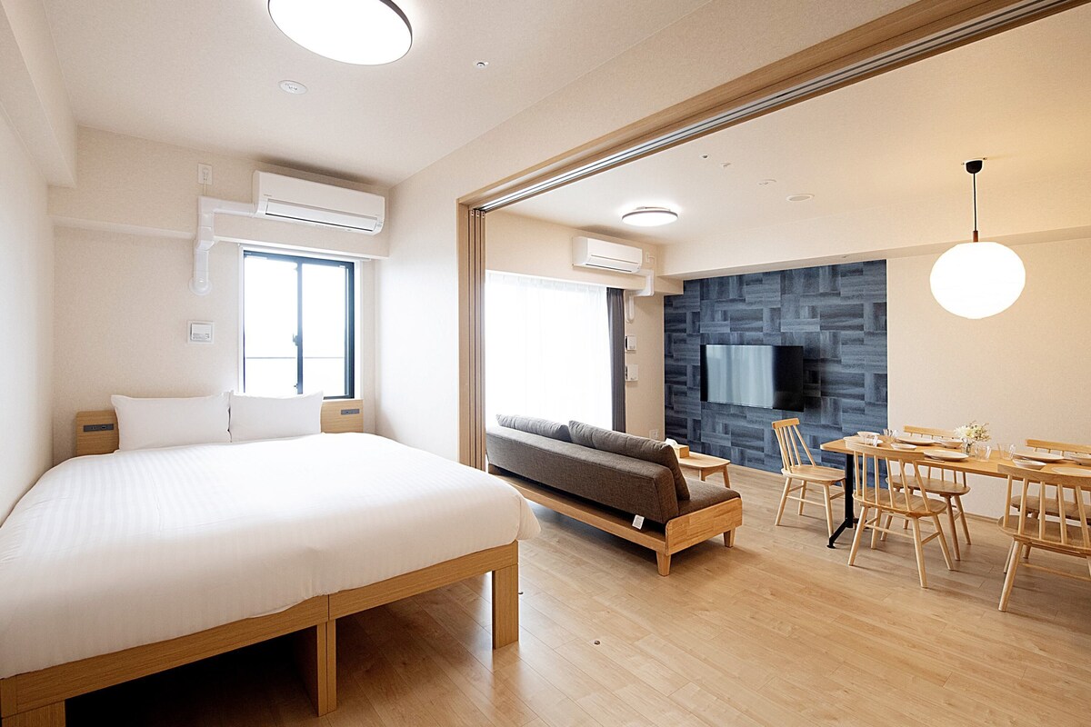 Habitació d'hotel moderna amb un llit gran, un sofà, zona de menjador amb taula i cadires, i televisió penjada a la pared.