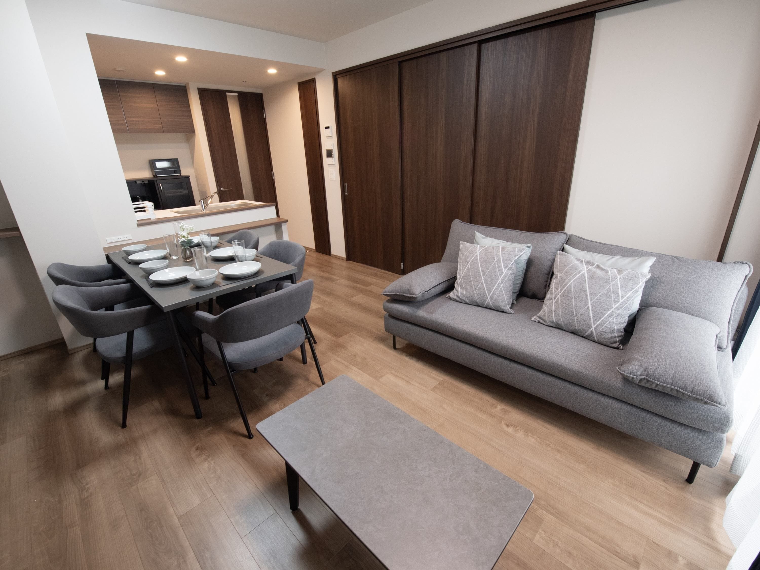 Una sala de estar moderna con un sofá gris, una mesa de comedor con sillas y una cocina al fondo.