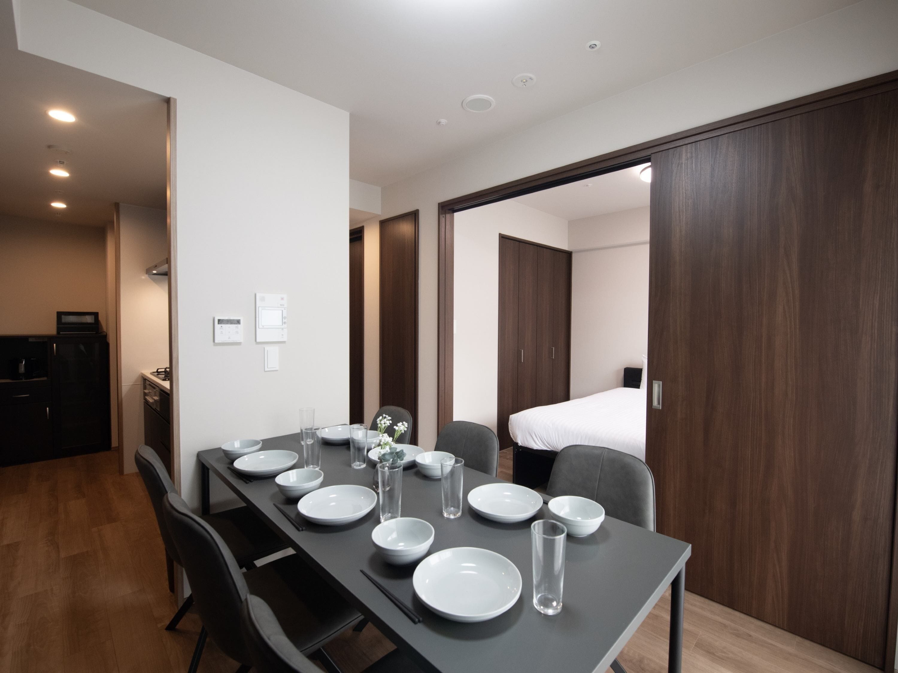 Une chambre d’hôtel moderne dotée d’un coin repas avec une longue table dressée pour quatre personnes, d’une kitchenette, d’une armoire et d’un lit dans la pièce adjacente.