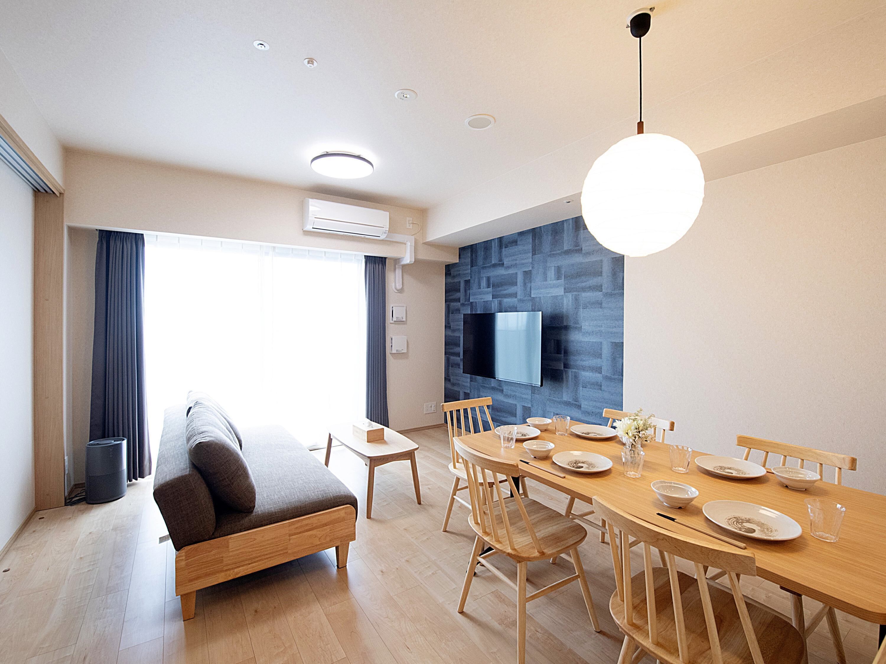 A modern living room with a sofa, dining table, and chairs. There is a flat-screen TV mounted on the wall and an air conditioning unit above the sofa.