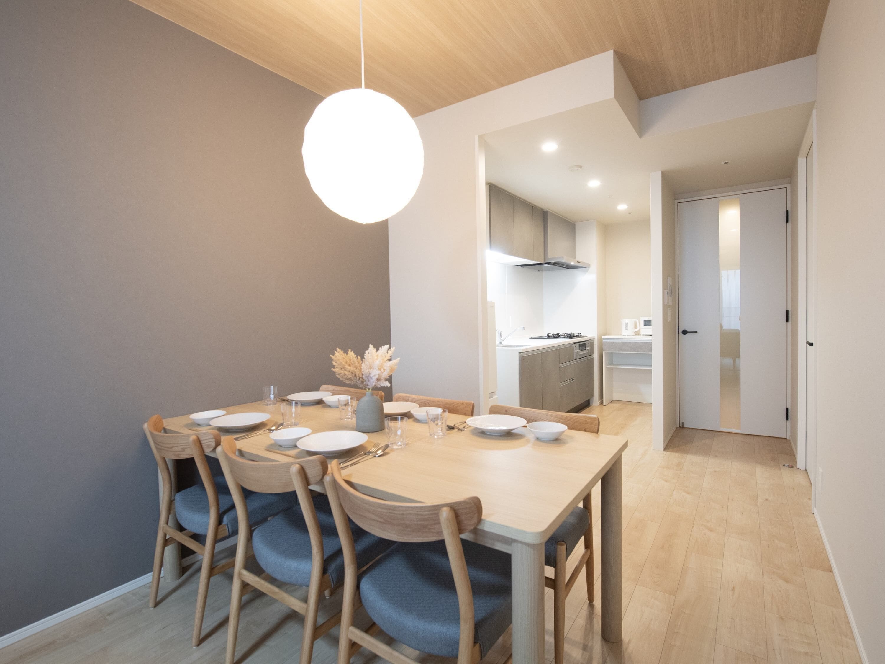 A dining area with a wooden table and chairs, a vase with dried flowers, and a hanging light fixture.