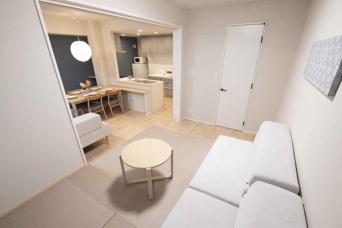 Un saló modern i minimalista amb un sofà, una taula rodona petita i una cuina visible a través d'una porta oberta.