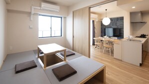 Een moderne, minimalistische kamer met tatami matten, een houten tafel en een keukenruimte zichtbaar door een open deur.