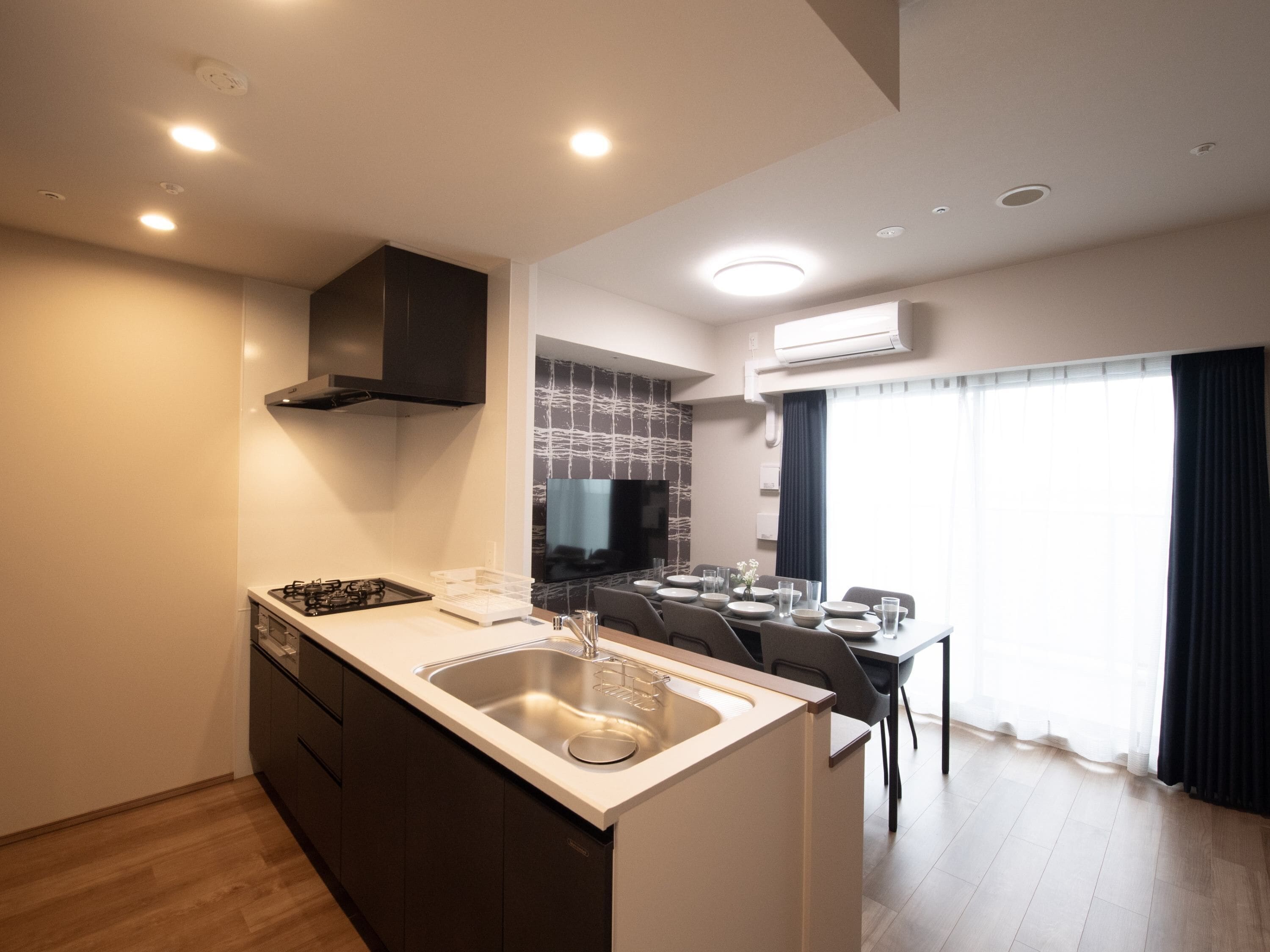 Una cocina moderna con estufa integrada, lavaplatos y armarios negros. Hay un comedor con mesa y sillas, y un ventanal con cortinas.
