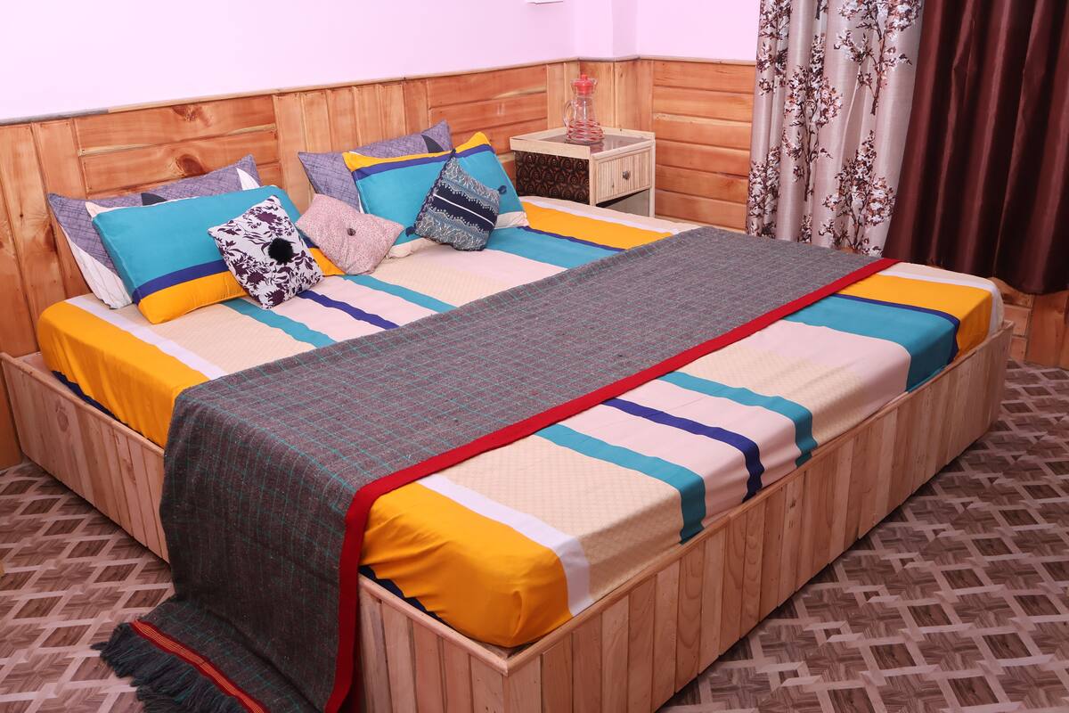 A wooden bed with a colorful bedspread and pillows, a small bedside table with a bottle, and patterned curtains.