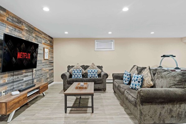 A living room with a large flat-screen TV mounted on a wooden wall, two sofas with patterned cushions, a coffee table, and a ceiling fan.