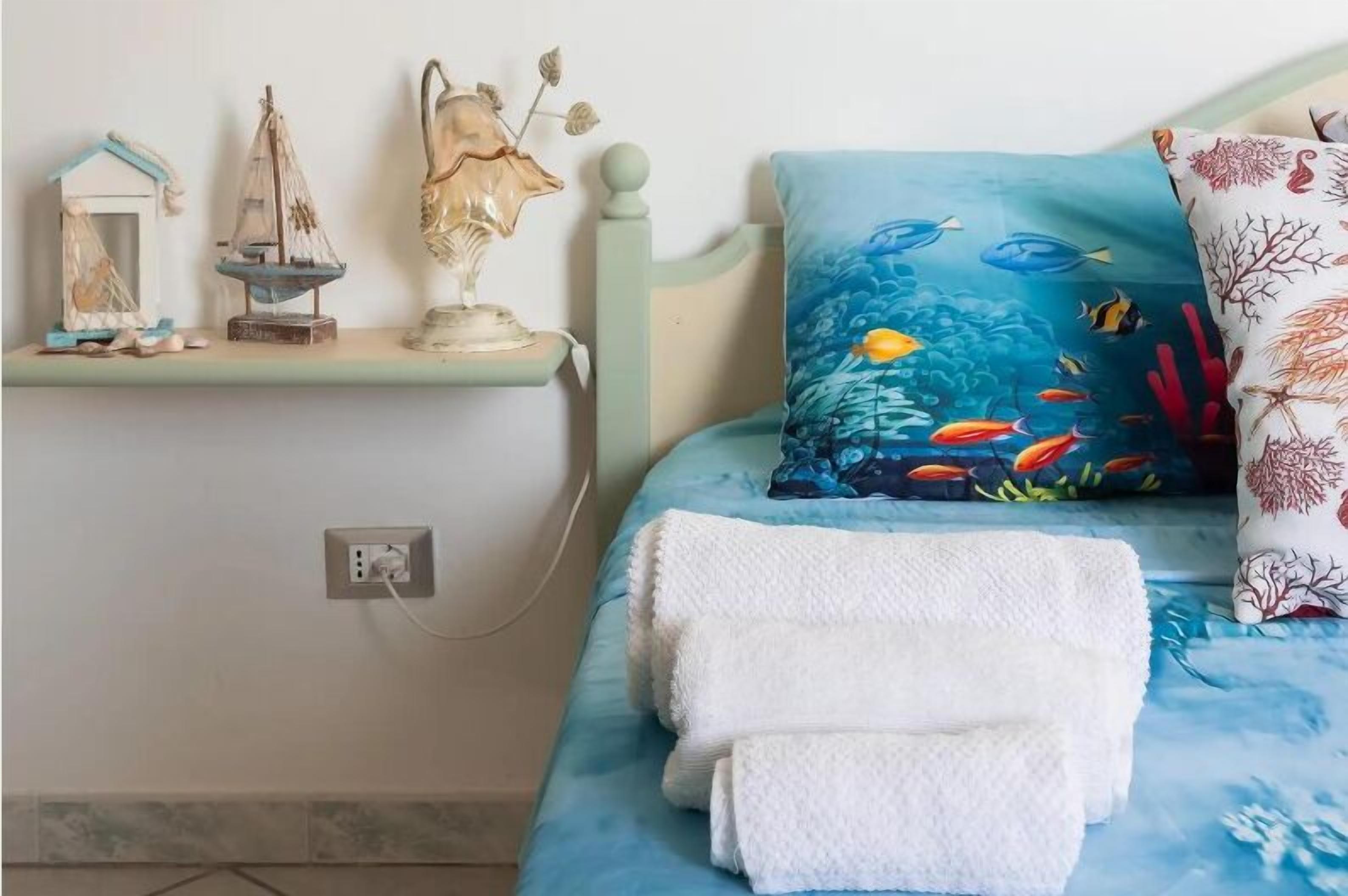 A bed with a blue ocean-themed pillow, a white towel, and decorative items on a shelf.