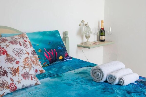 Una cama con una colcha de motivos marinos azules, un cojín con estampado de coral y una estantería con una botella de champán y copas.