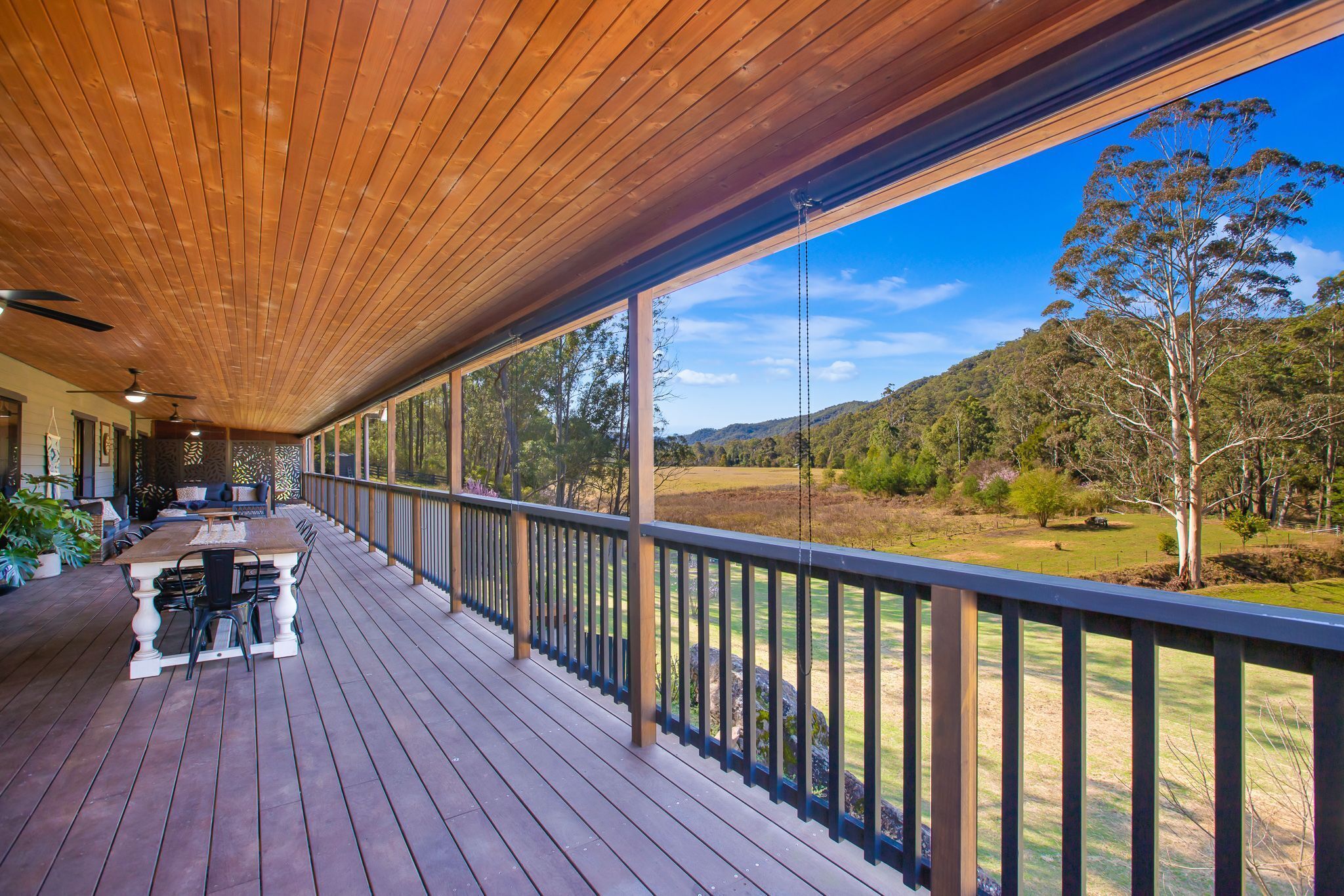 A wooden deck with a table and chairs, overlooking a grassy landscape with trees and hills.
