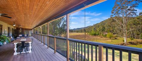A wooden deck with a table and chairs, overlooking a grassy landscape with trees and hills.