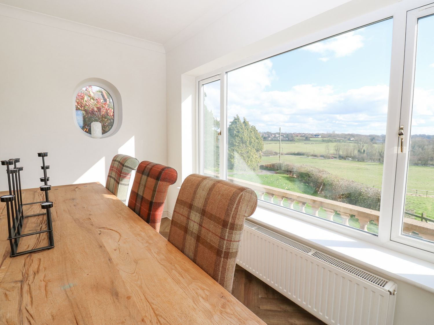 A dining area with a wooden table, plaid chairs, a round mirror, and a large window with a view of a green landscape.