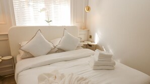 A neatly made bed with white linens and a white bathrobe on top, a bedside table with a lamp, and a window with blinds and a plant. - Arbour House Farm Bungalow (Durham)