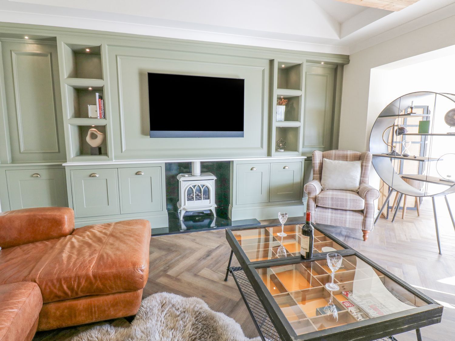 A living room with a large flat-screen TV mounted on a light green wall, a fireplace below, and a cozy seating area with a leather sofa and a plaid armchair.